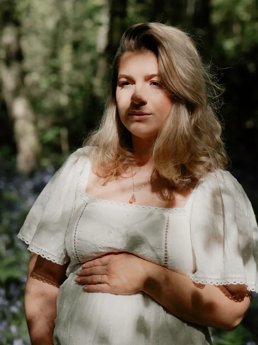 A pregnant woman with long light brown hair wearing a cream linen dress with puffed sleeves, standing outdoors with greenery behind her