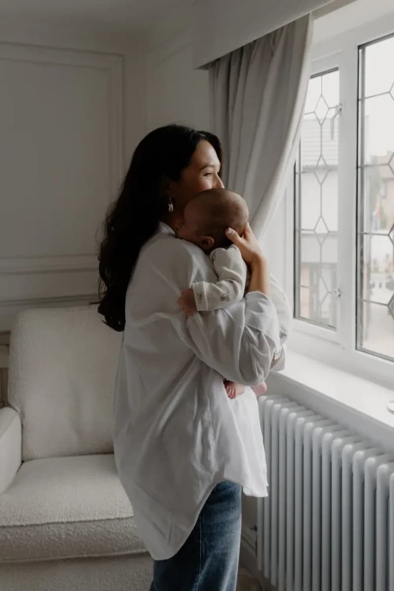 Woman in white cardigan holding a baby against her shoulder, looking out of a window with grey curtains